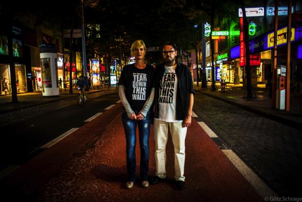 Zwei Personen stehen nachts auf einer belebten Straße und tragen T-Shirts mit der Aufschrift „Tear Down This Wall!“.