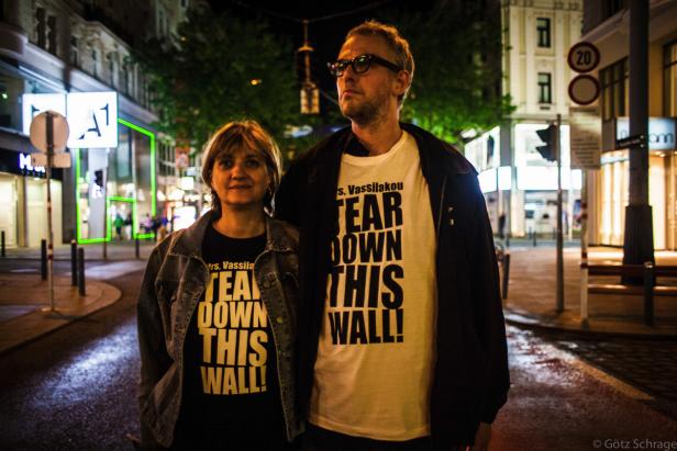 Zwei Personen stehen nachts in der Stadt und tragen T-Shirts mit der Aufschrift „Tear Down This Wall!“.