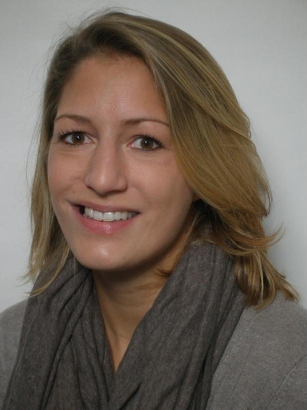Eine lächelnde Frau mit blonden Haaren und einem grauen Schal vor einem weißen Hintergrund.