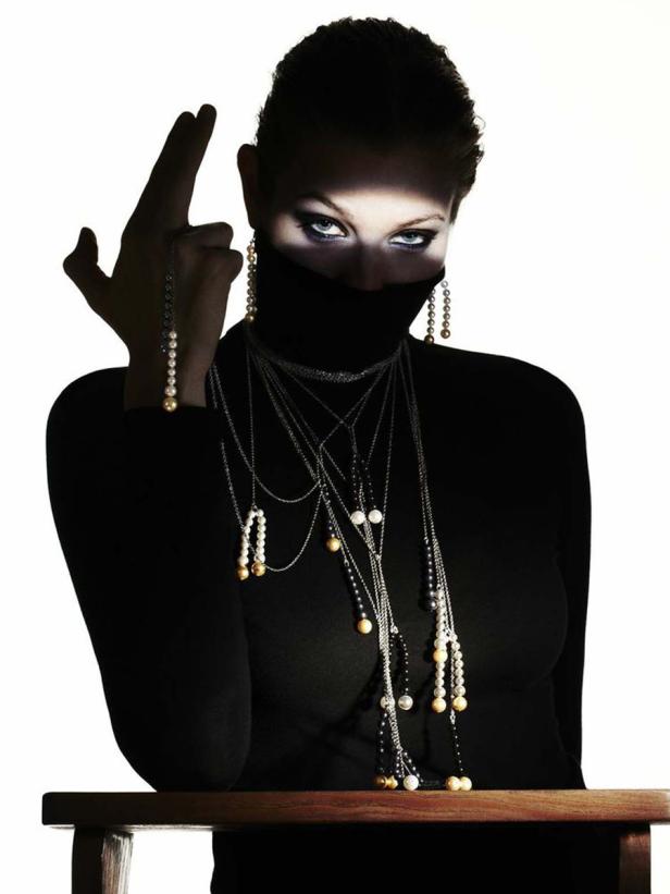 Eine Frau mit hochgeschlossenem, schwarzem Oberteil und vielen Ketten posiert vor einem hellen Hintergrund.