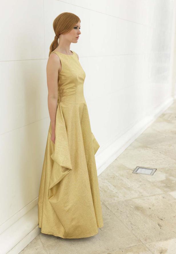 Eine Frau in einem goldenen, bodenlangen Kleid steht vor einer weißen Wand.