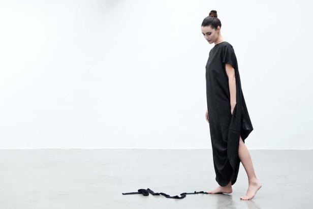 Eine Frau in einem schwarzen Kleid steht barfuß in einem minimalistischen Raum.