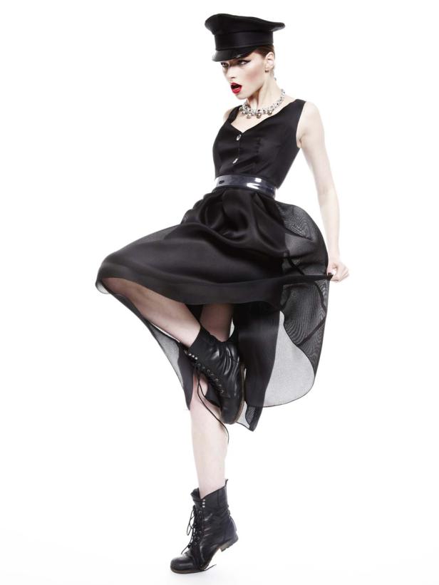 Eine Frau in einem schwarzen Kleid, Stiefeln und einer Mütze posiert vor einem weißen Hintergrund.