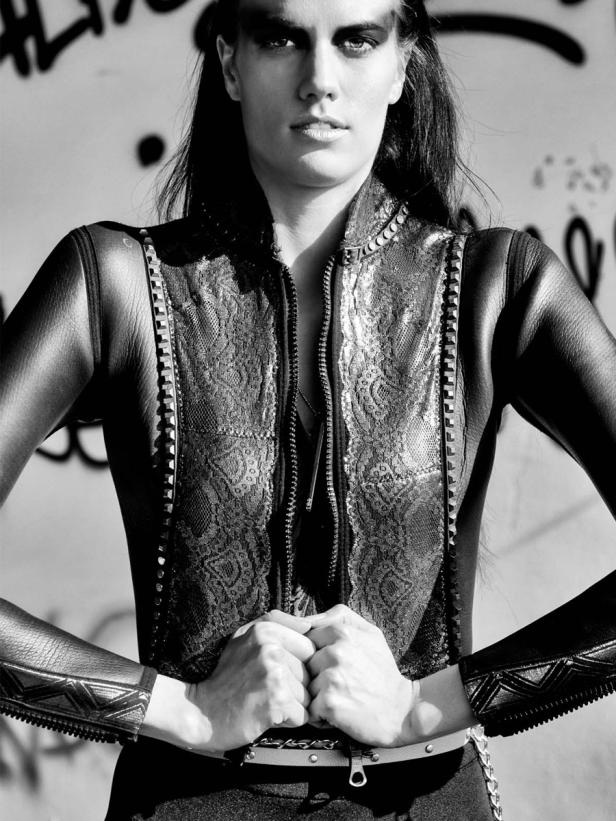 Eine Frau in einer Lederjacke mit Nietendetails posiert vor einer Wand mit Graffiti.