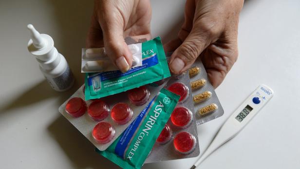 Eine Hand hält verschiedene Medikamente, darunter Aspirin Complex und ein Thermometer mit 38,8 Grad.