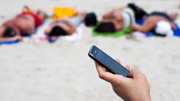Eine Hand hält ein Samsung-Smartphone am Strand, im Hintergrund liegen mehrere Personen.