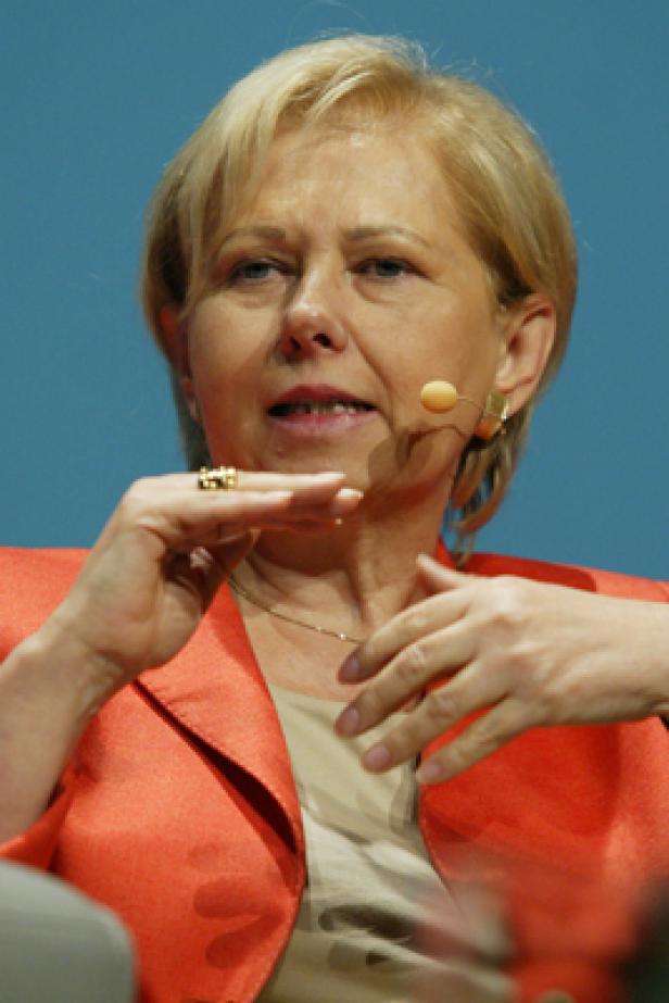 Eine Frau mit blonden Haaren und einem orangefarbenen Blazer spricht in ein Mikrofon.
