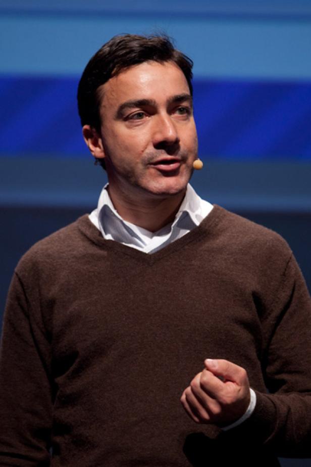 Ein Mann mit braunem Pullover spricht vor einem blauen Hintergrund.