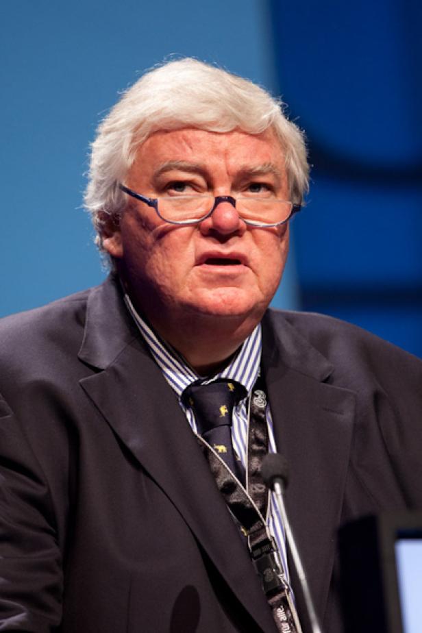Ein Mann mit Brille und Anzug spricht vor einem blauen Hintergrund.