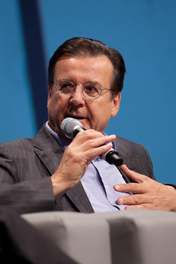 Ein Mann mit Brille spricht in ein Mikrofon vor einem blauen Hintergrund.