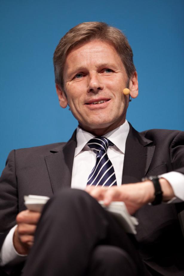 Ein Mann im Anzug mit Krawatte und Mikrofon sitzt vor einem blauen Hintergrund.