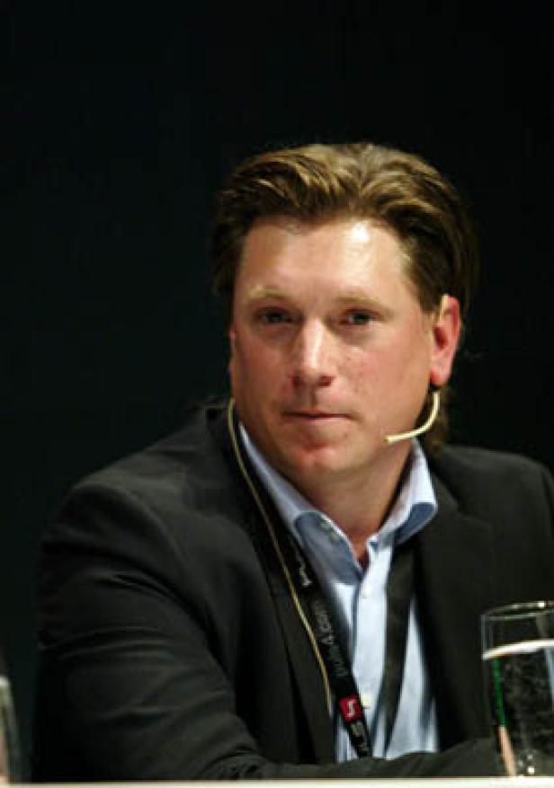 Ein Mann mit blonden Haaren und einem schwarzen Sakko spricht in ein Mikrofon.