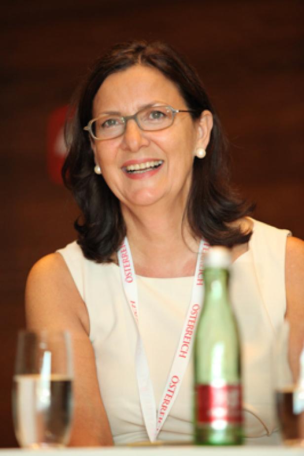 Eine lächelnde Frau mit Brille und einem „Österreich“-Band um den Hals sitzt an einem Tisch.