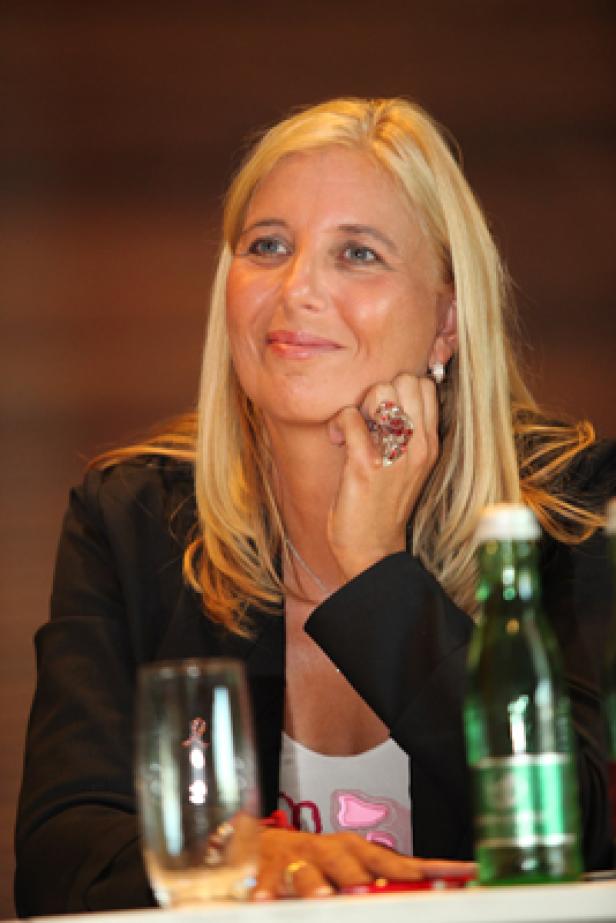 Eine blonde Frau mit Blazer und auffälligem Ring blickt aufmerksam zur Seite.