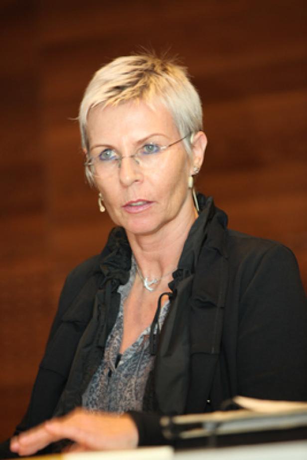 Eine Frau mit kurzen blonden Haaren und Brille spricht vor einem holzvertäfelten Hintergrund.
