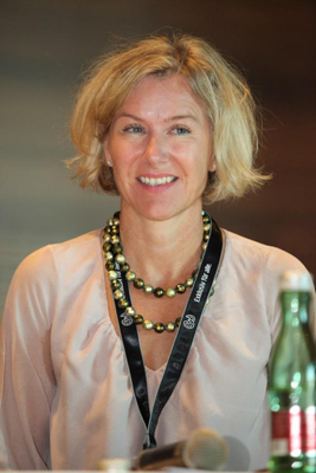 Eine Frau mit blonden Haaren und einer Perlenkette lächelt.