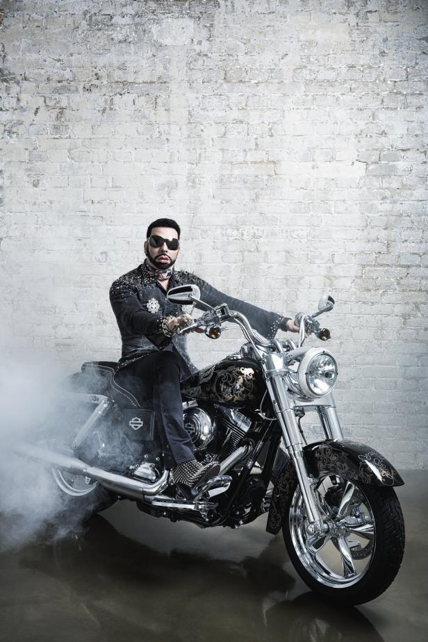 Ein Mann mit Sonnenbrille sitzt auf einem dekorierten Harley-Davidson-Motorrad vor einer Ziegelwand.