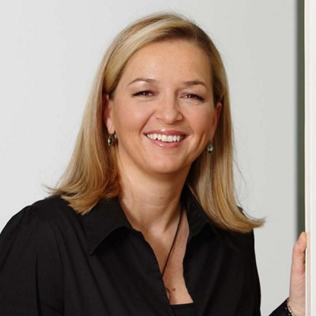 Eine lächelnde Frau mit blonden Haaren und schwarzem Hemd vor einem hellen Hintergrund.