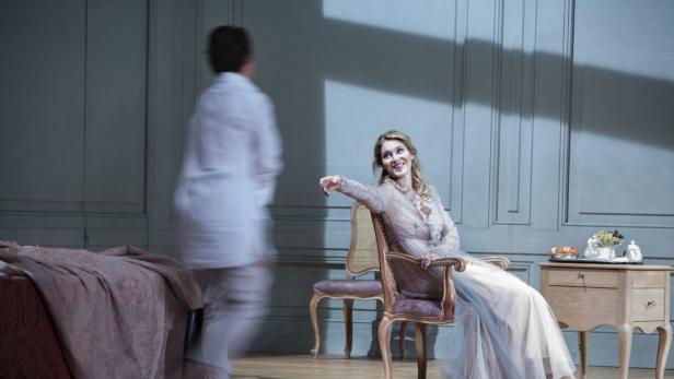 Eine Frau sitzt in einem eleganten Kleid auf einem Stuhl, während eine andere Person im Hintergrund vorbeigeht.