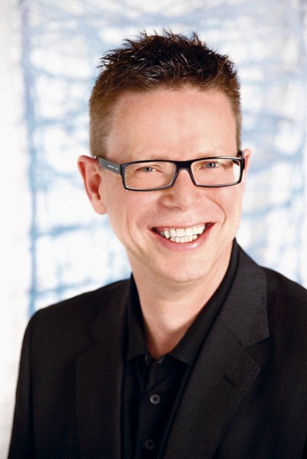 Ein lächelnder Mann mit Brille und dunklem Anzug vor einem hellen Hintergrund.