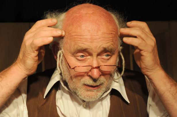 Ein älterer Mann mit Brille und Weste berührt seinen Kopf.