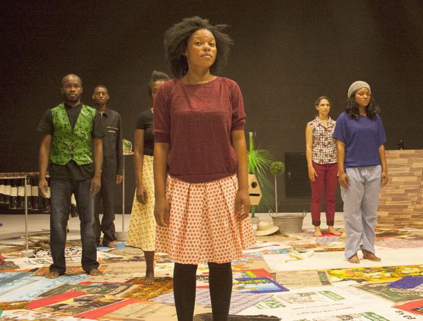 Mehrere Darsteller stehen barfuß auf einer mit Zeitungen bedeckten Bühne.