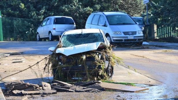 Ein beschädigtes Auto ist mit Schlamm und Pflanzenresten bedeckt; die Straße ist überflutet.