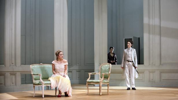 Eine Frau sitzt auf einem Stuhl, während zwei weitere Personen im Hintergrund stehen.