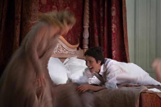 Eine Frau in einem weißen Hemd lacht, während eine andere Frau aufs Bett springt.