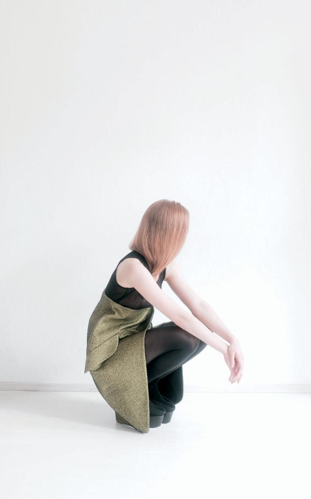 Eine Frau mit rotem Haar kauert in einem goldenen Kleid und schwarzen Strumpfhosen vor einer weißen Wand.