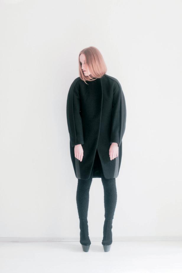 Eine Frau mit roten Haaren trägt einen schwarzen Mantel, Leggings und Stiefel.