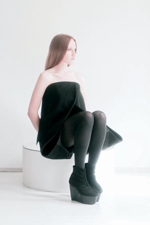 Eine Frau in einem schwarzen, trägerlosen Kleid und schwarzen Stiefeln sitzt auf einem weißen Sockel.