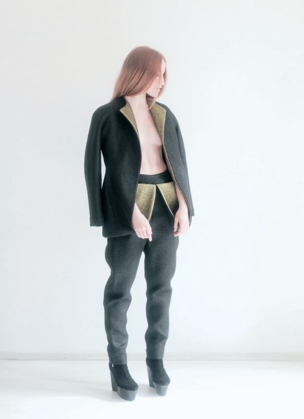 Eine Frau mit langen, rötlichen Haaren trägt einen schwarzen Hosenanzug mit goldenen Akzenten und Plateauschuhen.