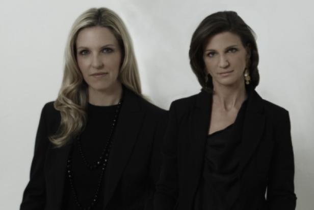 Zwei Frauen in schwarzen Blazern posieren vor einem hellen Hintergrund.