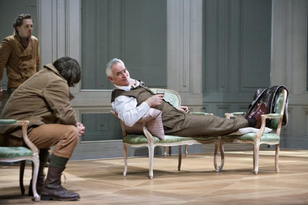 Ein Mann liegt entspannt in einem Sessel, während zwei andere Männer im Hintergrund stehen.