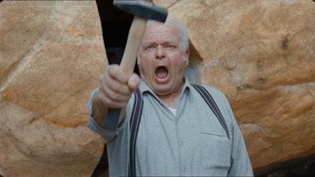 Ein Mann mit Hosenträgern hält einen Hammer in der Hand und schreit.