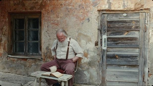 Ein älterer Mann mit Bart sitzt vor einem Haus und liest ein Buch.