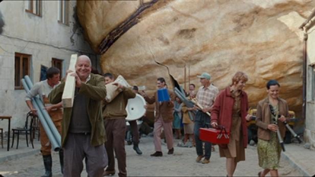 Eine Gruppe von Menschen trägt Baumaterialien durch eine Gasse vor einem großen Felsen.