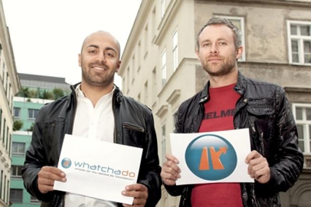 Zwei Männer posieren mit Schildern der Firma „whatchadoo“ in einer städtischen Umgebung.