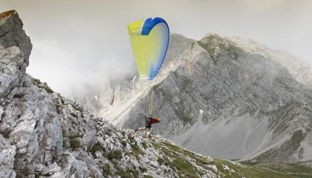 Ein Mann startet mit einem gelb-blauen Gleitschirm von einem felsigen Berghang.