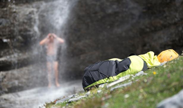 Ein Mann duscht unter einem Wasserfall, während Ausrüstung daneben liegt.