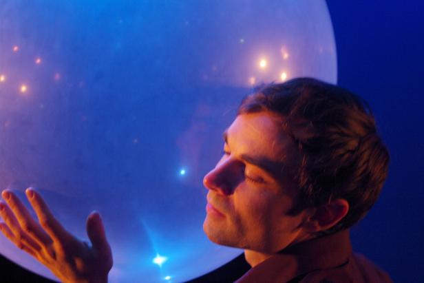 Ein Mann steht vor einer großen, leuchtenden Kugel mit geschlossenen Augen.