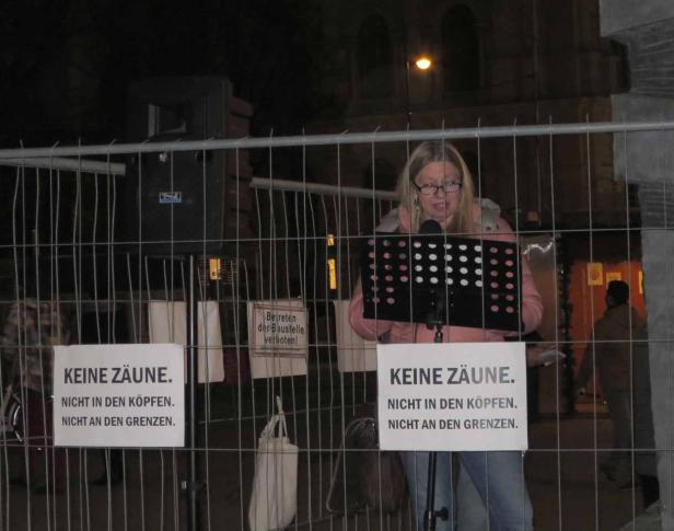 Eine Frau spricht bei einer Kundgebung hinter einem Bauzaun, auf dem Schilder mit der Aufschrift „Keine Zäune. Nicht in den Köpfen. Nicht an den Grenzen“ hängen.