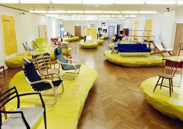 In einem Ausstellungsraum sind verschiedene Stühle und Sitzmöbel auf gelben Podesten ausgestellt.