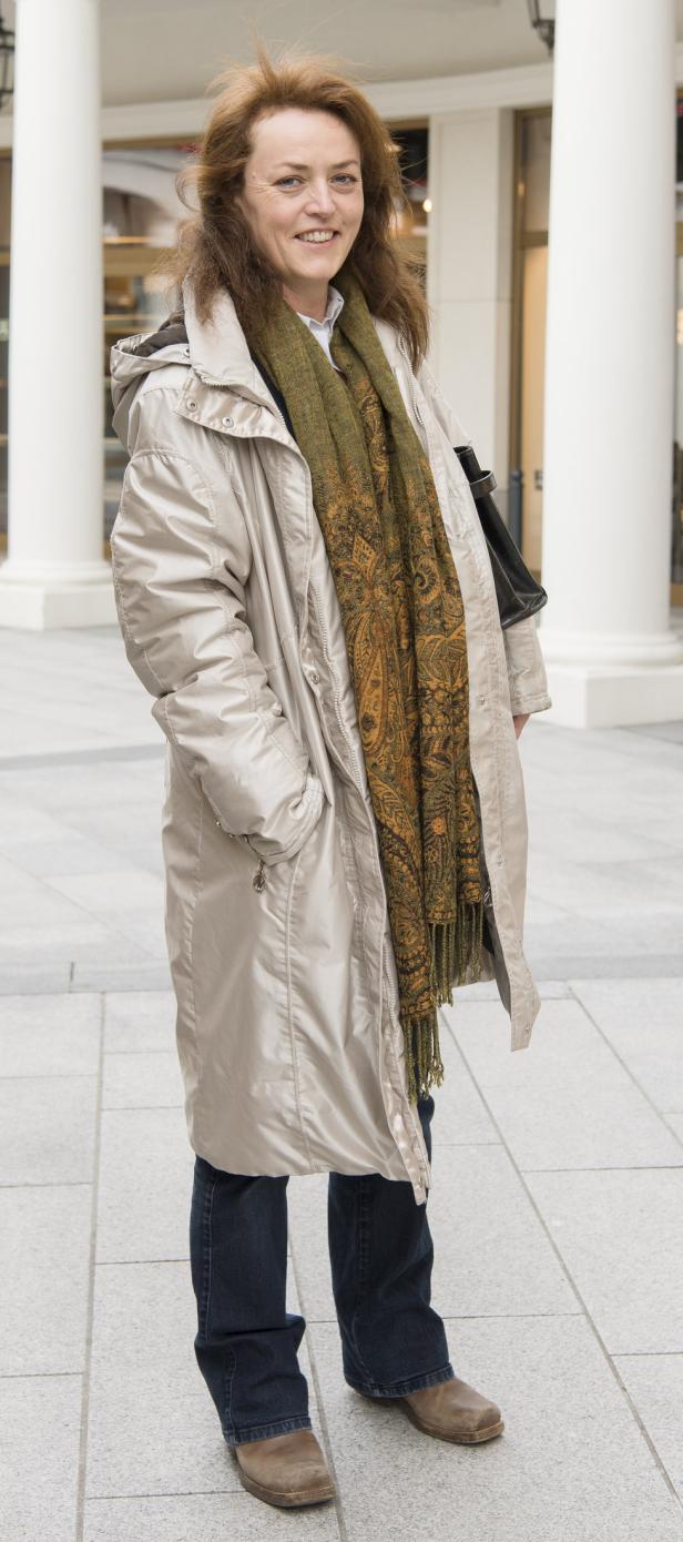 Eine Frau mit braunen Haaren, bekleidet mit einem beigen Mantel und einem gemusterten Schal.
