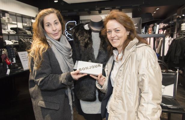 Zwei Frauen präsentieren in einem Bekleidungsgeschäft eine Handtasche mit der Aufschrift „Bonjour“.