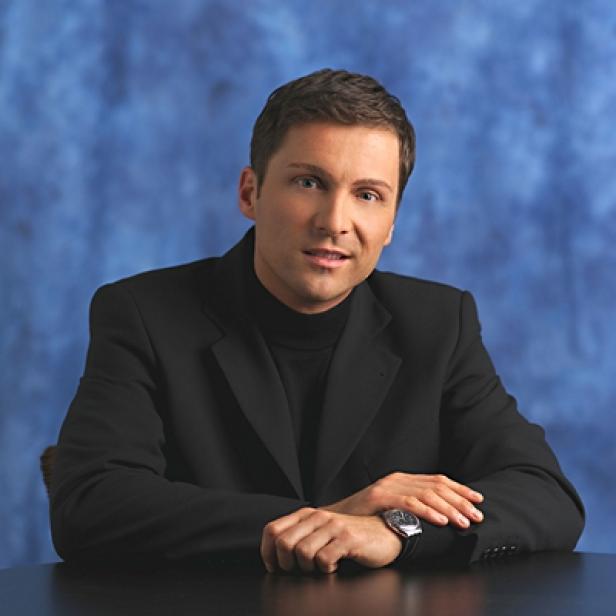 Ein Mann in einem schwarzen Anzug und Rollkragenpullover sitzt an einem Tisch.
