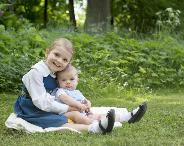 Ein älteres Mädchen im Dirndl hält ein Baby auf einer Wiese.