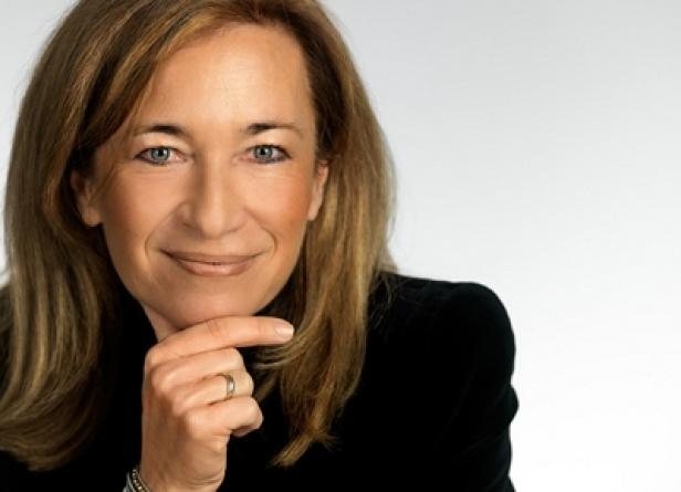 Eine Frau mit blonden Haaren und einem schwarzen Blazer lächelt in die Kamera.