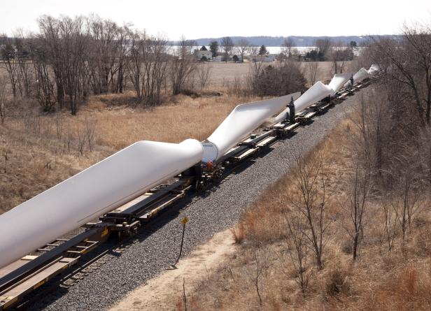 Ein Zug transportiert Windkraftanlagenflügel durch eine ländliche Gegend.